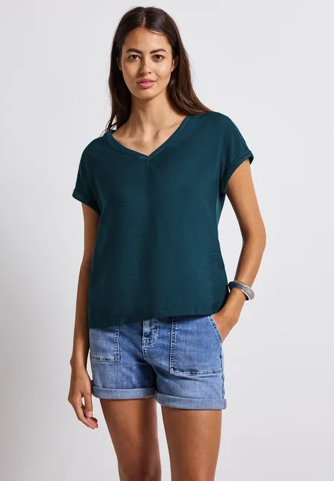 Frau mit dunkelgrünem Shirt und Jeansshorts, steht lächelnd vor weißem Hintergrund.